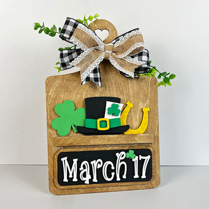 Janet's Craft Corner offers a DIY interchangeable home decor kit featuring a breadboard base and insert decorated with "March 17," a black top hat with a green band, green shamrock, gold horseshoe, plaid ribbon with lace trim, and green plant accents for charming festive decor.