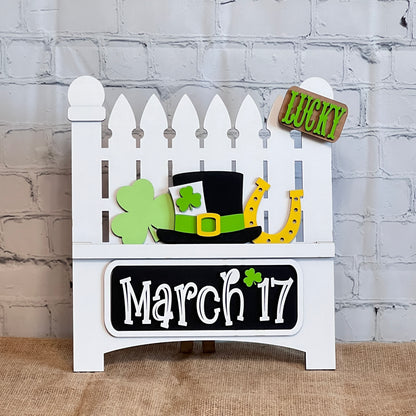 Janet's Craft Corner offers a DIY Garden Fence decor kit, featuring a white picket fence with St. Patrick's Day elements: a green shamrock, black top hat, yellow horseshoe, and "March 17" in white. An interchangeable "Lucky" sign completes the set.