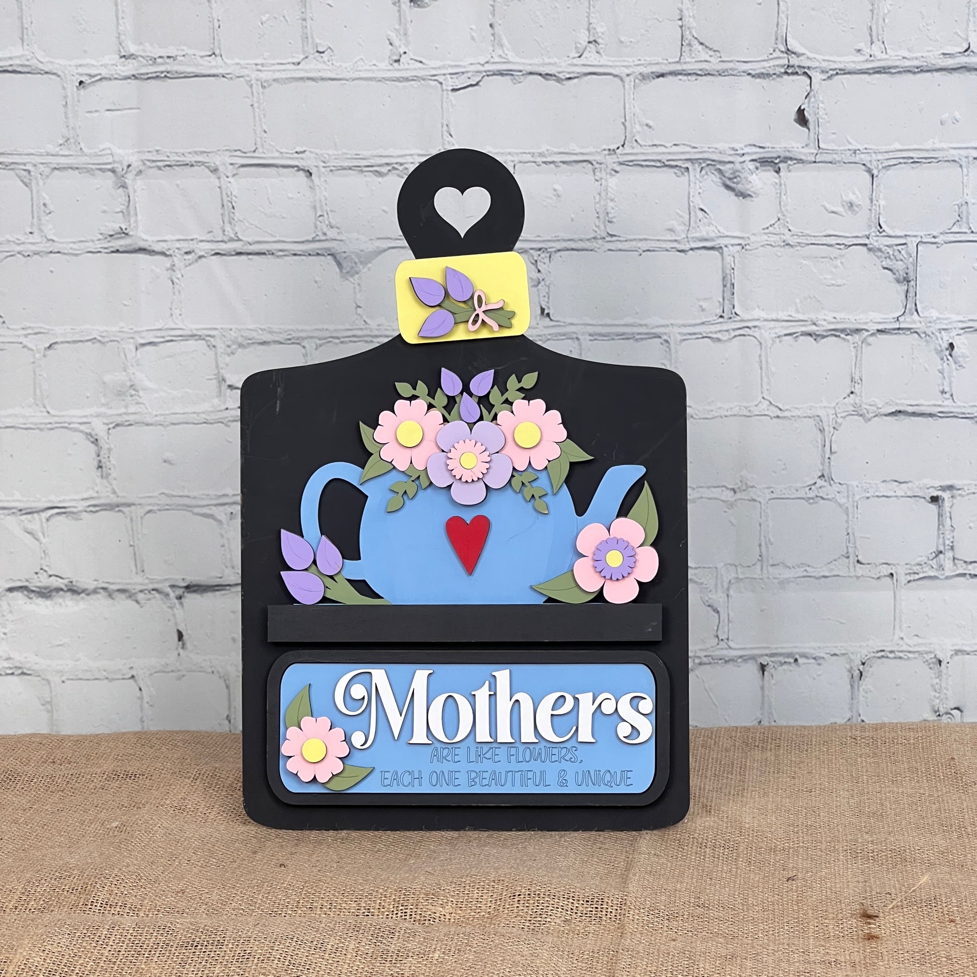 Janet's Craft Corner offers a DIY interchangeable home decor kit featuring a breadboard base and an insert with a teapot brimming with pink flowers. The accompanying message, "Mothers are like flowers, each one beautiful & unique," is adorned with heart and flower motifs. Perfect for personalization.