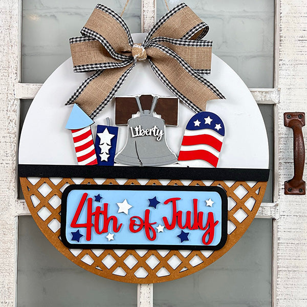 Janet's Craft Corner offers a DIY kit for 4th of July décor. It includes a round wooden sign with Liberty Bell, American flags, rockets cutouts, and a checkered bow. "4th of July" is highlighted in red on a starry blue-and-white background, perfect as an interchangeable door hanger.