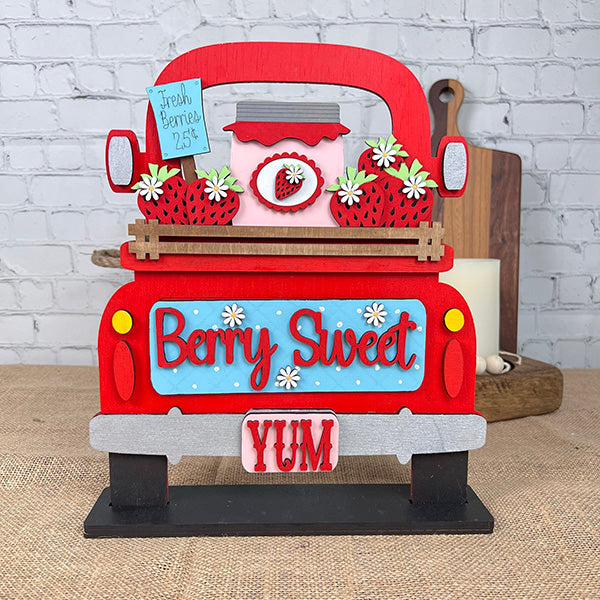 Janet's Craft Corner offers a DIY interchangeable home decor kit featuring an antique truck base filled with strawberries and a jam jar. The backboard reads "Berry Sweet" and "YUM" in colorful letters, set against a light brick wall and wooden tabletop for charming decor.