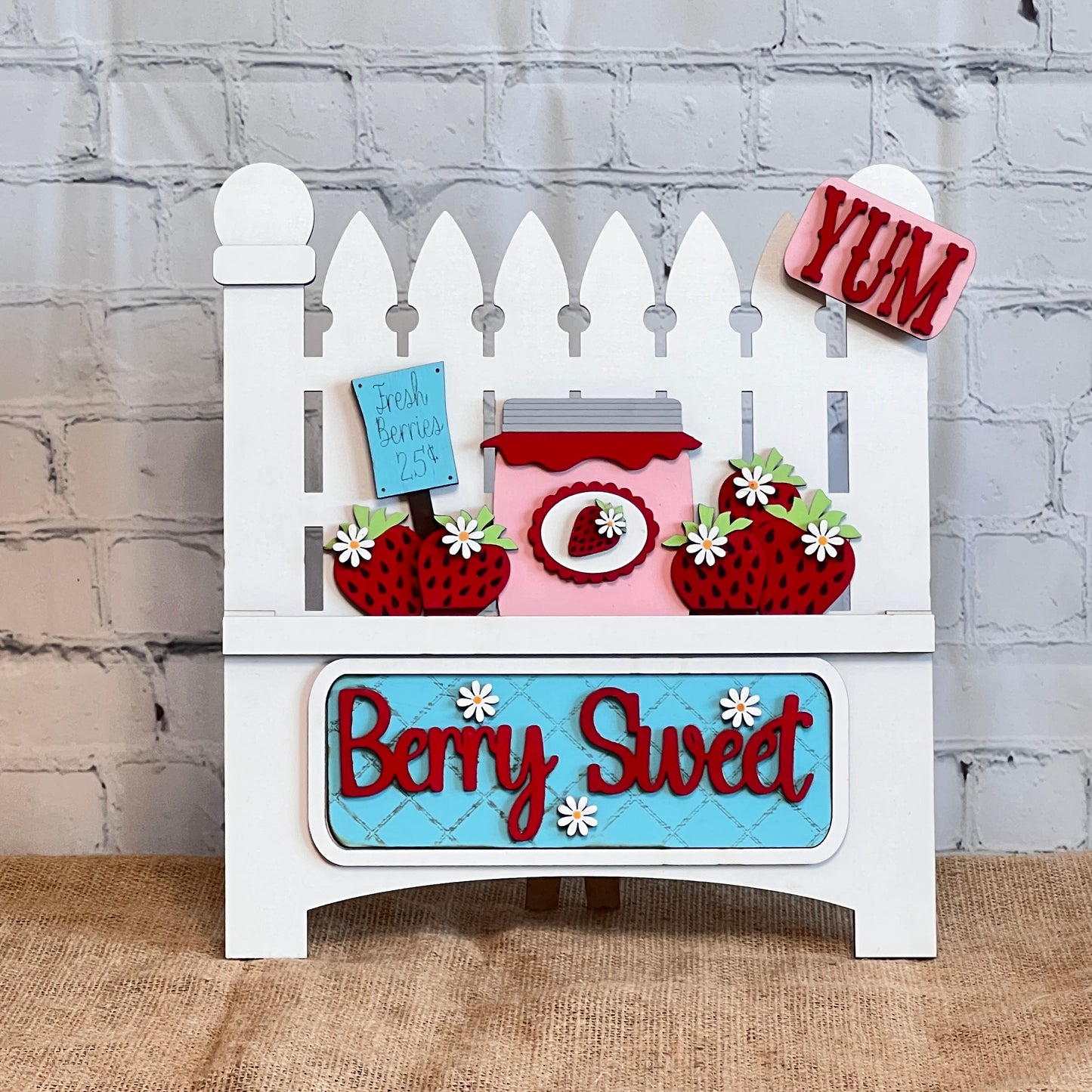 Janet's Craft Corner offers a Garden Fence with Insert DIY kit, featuring a white picket fence design. It includes an interchangeable insert with a pink jar and red lid surrounded by strawberries, "Berry Sweet" text on blue, a "Fresh Berries 25¢" sign, and a red "YUM" tag on top.