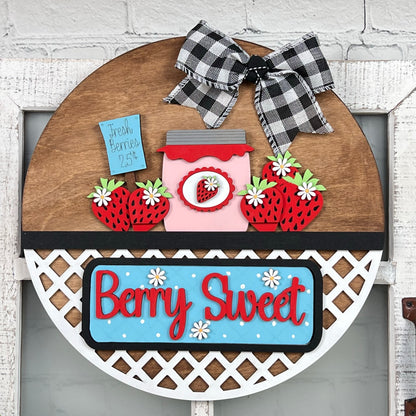 Janet's Craft Corner offers a delightful DIY home décor kit featuring a round wooden sign with strawberries, daisies, and a jar of jam. The blue sign reads "Fresh Berries 25¢" and "Berry Sweet," while a plaid bow tops it off, perfect as an interchangeable door hanger.