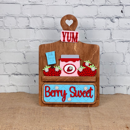 Janet's Craft Corner offers a Breadboard Interchangeable Base with Insert—ideal for home decor. It includes a decorative wooden board with a heart-shaped handle featuring "YUM," strawberries, jam illustration, and "Berry Sweet" on a blue plaque, set against a white brick wall background.