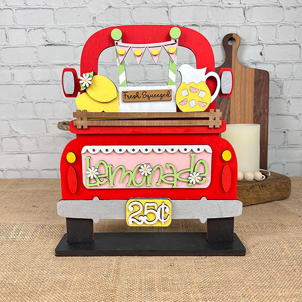 Janet's Craft Corner offers the Antique Truck Base with Insert DIY kit, featuring a hand-painted, colorful model of an antique truck with a "Fresh Squeezed" lemonade sign. It includes lemons, a pitcher, and a "Lemonade 25¢" sign against burlap and brick for charming interchangeable decor.