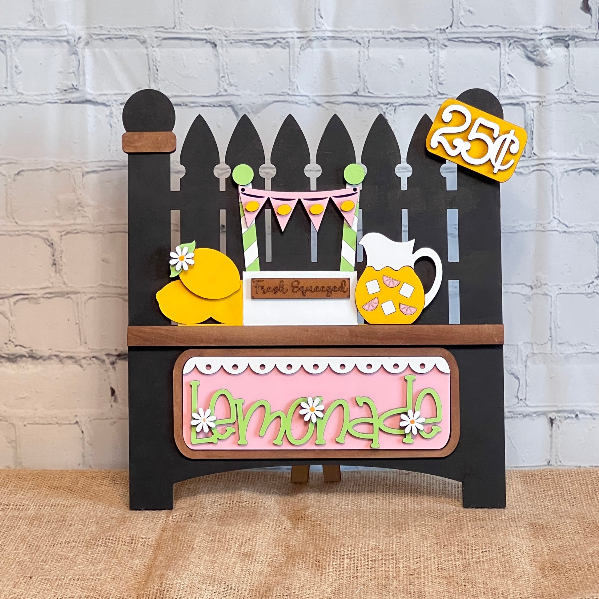 The black picket-fence-shaped sign from Janet's Craft Corner features a colorful lemonade theme with lemons, a pitcher, and "25¢" details. It includes playful green and white "lemonade" letters on a pink background. This DIY kit offers an interchangeable insert for creative home decor!.