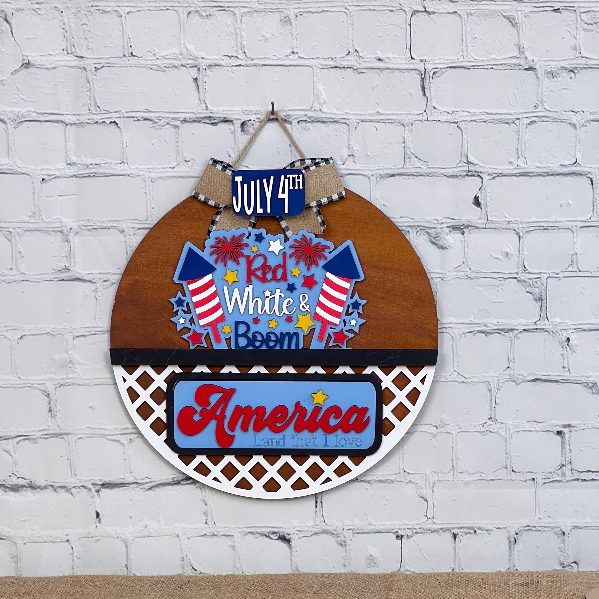 A door hanger from Janet's Craft Corner featuring fireworks, stars, and stripes on a white brick wall reads "July 4th, Red White & Boom." This interchangeable home décor kit blends wood and lattice textures, perfect for patriots and DIY enthusiasts alike.