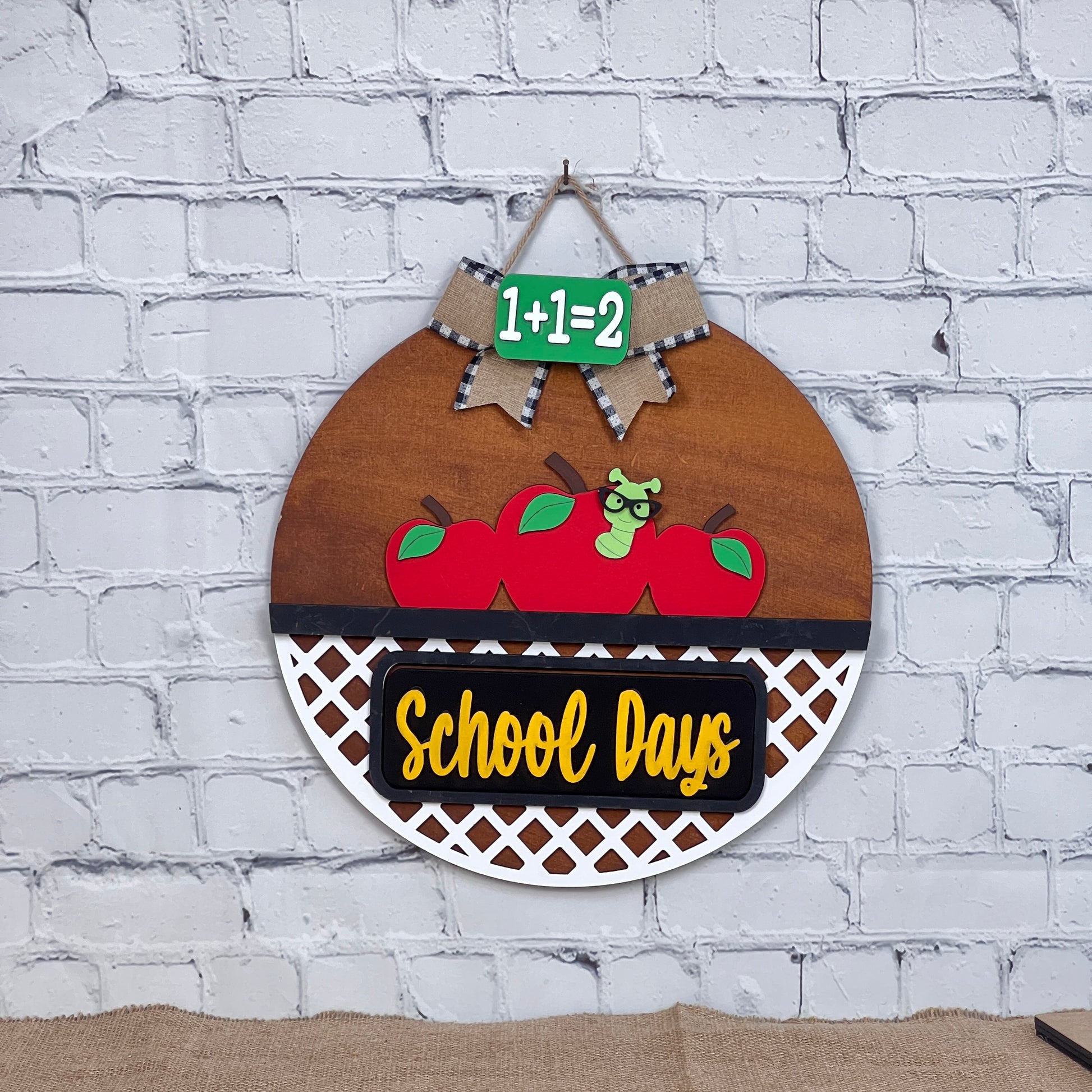 Janet's Craft Corner offers an interchangeable door hanger featuring three red apples, a glasses-wearing worm, and a "School Days" banner. A blackboard reading "1+1=2" with a bow adds charm. Perfect for DIY home décor.