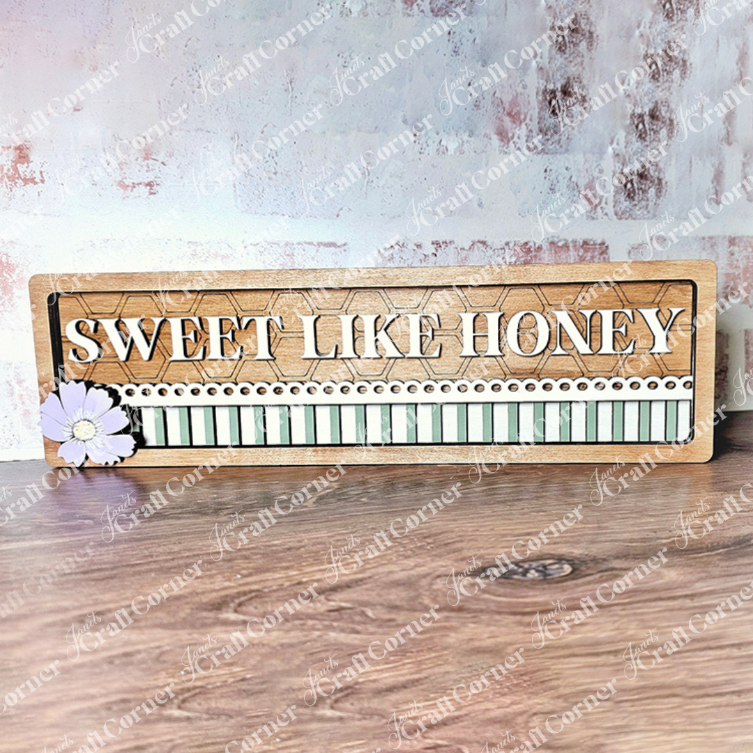 Janet's Craft Corner presents the Sweet Like Honey Chunky Shelf Sitter, a DIY wooden sign kit featuring bold "Sweet Like Honey" lettering. It includes black and white stripes with dots, accompanied by a purple flower on the left, set against a textured pink and gray wall background.