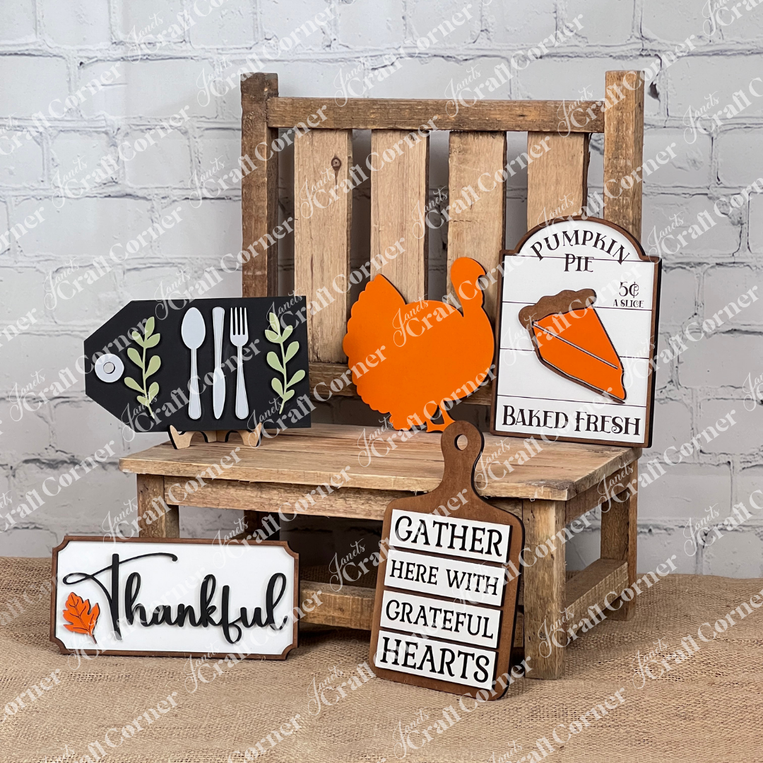 On a wooden chair, you'll find the Thankful Decor Set from Janet's Craft Corner, featuring five fall-themed signs: a turkey design, "Pumpkin Pie Baked Fresh," utensils on black, "Thankful," and "Gather Here with Grateful Hearts." This DIY home decor craft kit adds personalized charm with its leaf accents against a white brick wall.