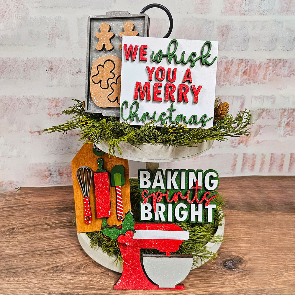 A festive display showcases a two-tiered stand adorned with greenery and gingerbread cookies, complemented by a sign that reads "We wish you a Merry Christmas." Below this, another sign proclaims "Baking Spirits Bright" next to a red mixer. Enhance your home décor with holiday cheer using Janet's Craft Corner's charming Baking Spirits Bright Decor Set - 5 Pieces - DIY Home Decor Kit!