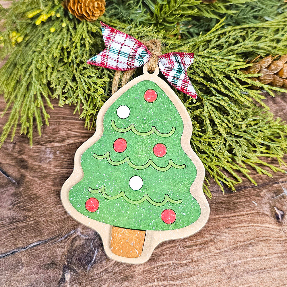 A wooden ornament from Janet's Craft Corner, part of the Sugar Cookie Ornaments - Set of 3, captures the delightful appearance of a sugar cookie. It shimmers with green glitter, red and white circles, and is topped with a ribbon in red, white, and green plaid. The ornament hangs gracefully against a backdrop of wood and lush green foliage.