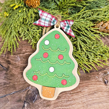 A wooden ornament from Janet's Craft Corner, part of the Sugar Cookie Ornaments - Set of 3, captures the delightful appearance of a sugar cookie. It shimmers with green glitter, red and white circles, and is topped with a ribbon in red, white, and green plaid. The ornament hangs gracefully against a backdrop of wood and lush green foliage.