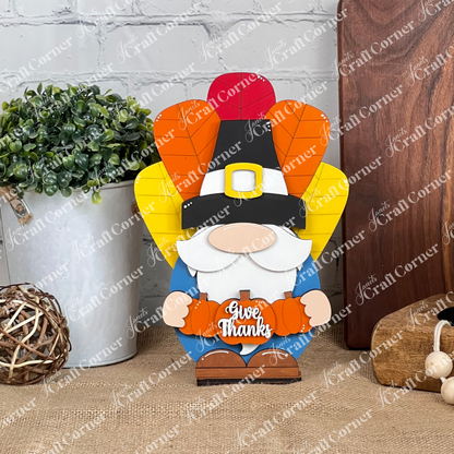 The Turkey Gnome Shelf Sitter from Janet's Craft Corner is a delightful DIY home decor craft kit featuring a Thanksgiving-themed gnome adorned with a pilgrim hat and holding a pumpkin inscribed with "Give Thanks." This charming piece is complemented by colorful, leaf-shaped feathers in the background, alongside an elegant potted plant and decorative wicker ball to enhance your home décor on any wooden surface.