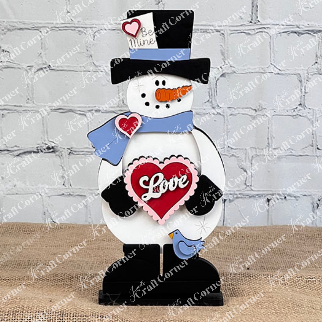 Janet's Craft Corner presents the Valentine Snowman Shelf Sitter: a charming decor piece featuring a "Love" heart, black top hat with "Be Mine" tag, and a blue bird on its foot. Perfect against any gray brick wall, this DIY seasonal craft kit is an ideal Valentine's Day decoration.