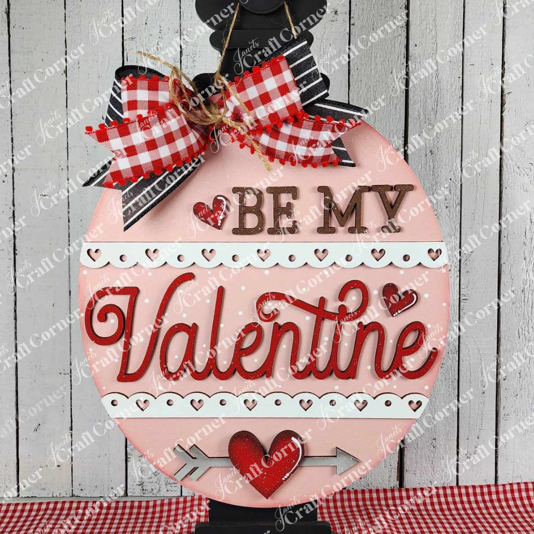 Janet's Craft Corner offers a DIY home décor craft kit: the Be My Valentine Door Hanger. This set features a pink round hanger with a red and white gingham bow, "Be My Valentine" cut-outs, red hearts, arrow designs, and a white wooden texture background for Valentine's Day enthusiasts.