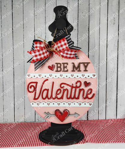 The "Be My Valentine" door hanger DIY craft kit by Janet's Craft Corner features a rustic wooden design with red and brown writing, hearts, and an arrow motif. It includes a red and white checkered bow for a hand-painted charm perfect for Valentine's seasonal décor.