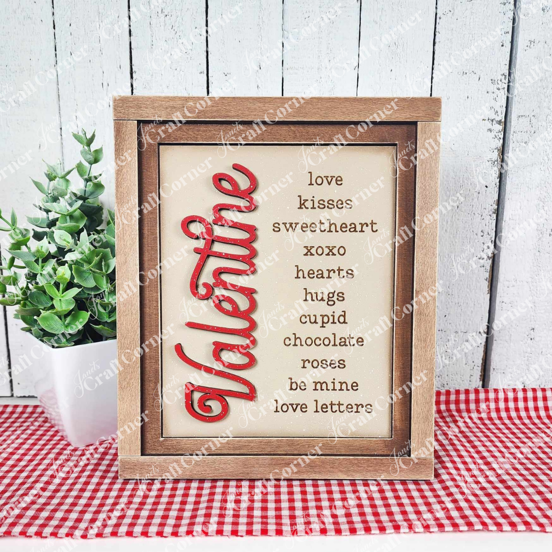 The Valentine Words Sign by Janet's Craft Corner features a wooden frame with "Valentine" in red vertical text and hand-painted words like "love," "kisses," and "sweetheart." This interchangeable decor piece is displayed on a red-and-white checkered cloth next to a lush potted plant.