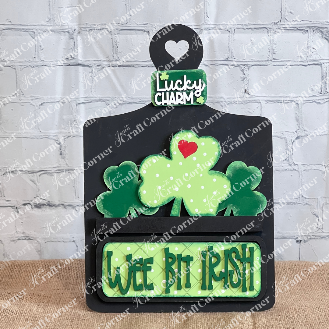 The Wee Bit Irish Interchangeable Insert by Janet's Craft Corner is a decorative wooden sign with a heart-topped handle, featuring green shamrocks and a "Wee Bit Irish" plaque. Perfect for any festive occasion, display it against a white brick wall to showcase your Irish pride.