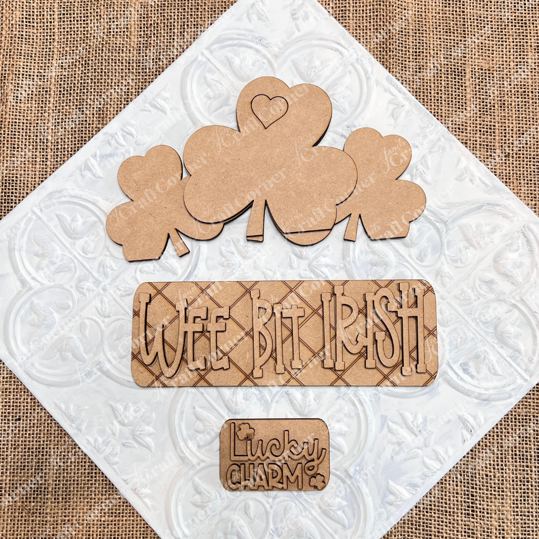 Janet's Craft Corner presents the "Wee Bit Irish" Interchangeable Insert: featuring wooden shamrocks, one with a heart, a plaque reading "WEE BIT IRISH," and a small piece reading "Lucky Charm" beside a shamrock. The burlap background enhances this charming DIY home decor kit.