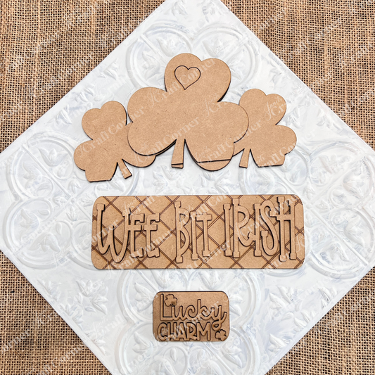 Janet's Craft Corner presents the "Wee Bit Irish" Interchangeable Insert: featuring wooden shamrocks, one with a heart, a plaque reading "WEE BIT IRISH," and a small piece reading "Lucky Charm" beside a shamrock. The burlap background enhances this charming DIY home decor kit.