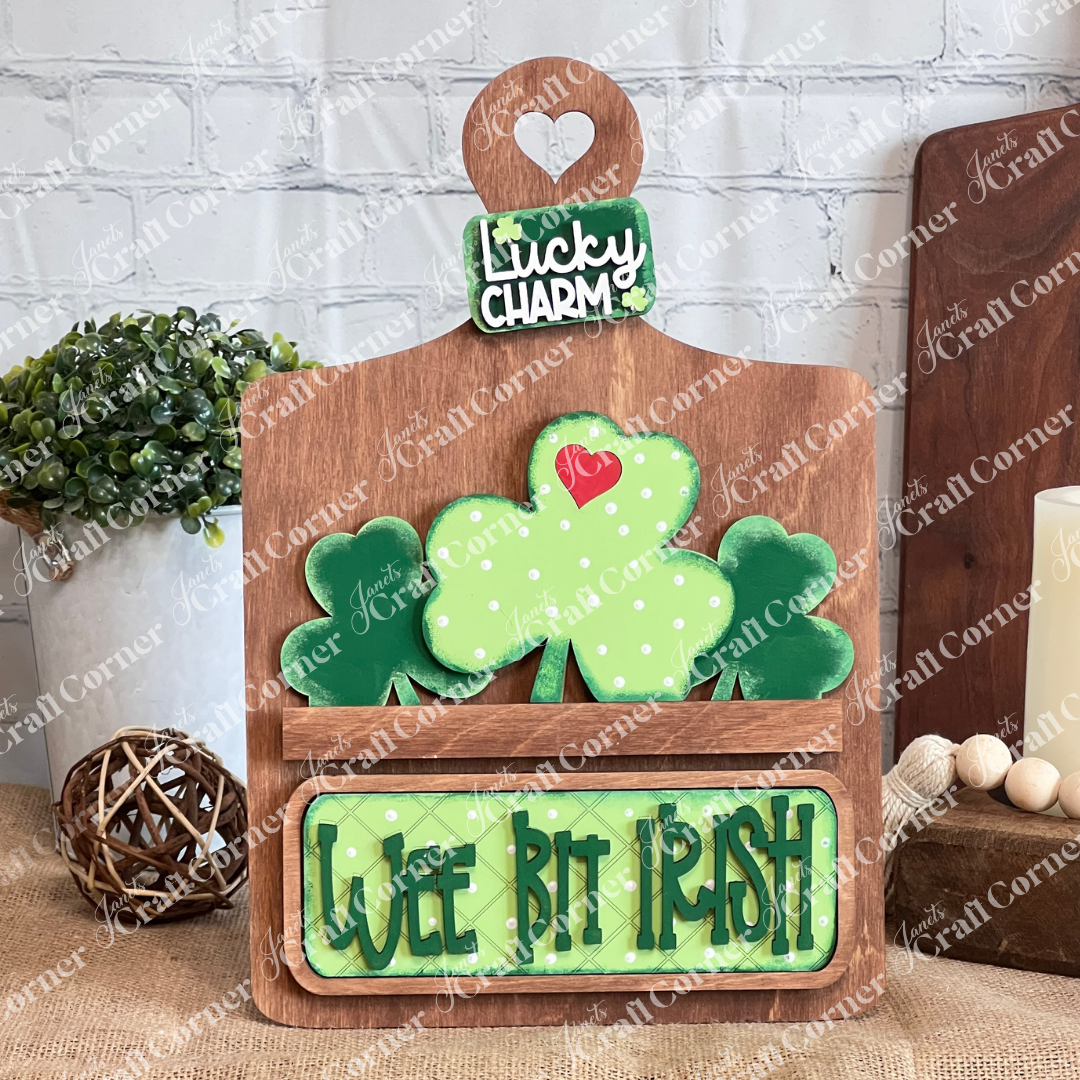 The "Wee Bit Irish Interchangeable Insert" by Janet's Craft Corner features a wooden piece with a heart cutout adorned with polka-dot shamrocks and a felt heart, topped by a "Lucky Charm" sign. It's perfect for themed items, celebrating Irish pride with added charm from a background plant.