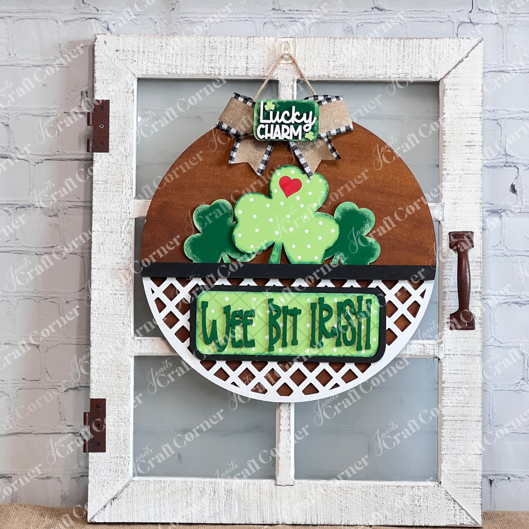 The "Wee Bit Irish" Interchangeable Insert by Janet's Craft Corner showcases Irish pride with a shamrock, hearts, and a "Lucky Charm" label on rustic white lattice. Ideal as a DIY craft kit, it elegantly hangs in a white frame against a brick wall.