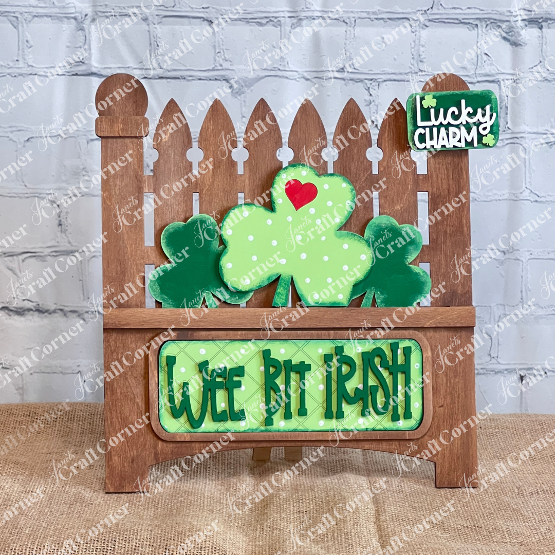 The "Wee Bit Irish Interchangeable Insert" by Janet's Craft Corner features a wooden picket fence sign with a green shamrock, red heart, and "Lucky Charm" text. A green plaque reads "Wee Bit Irish," set against a wood-textured background, making it an ideal DIY home decor craft kit.