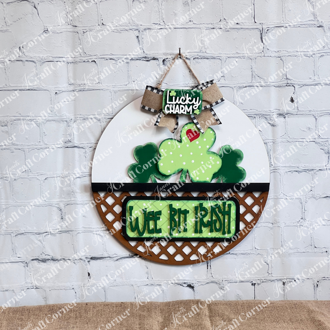 The Wee Bit Irish Interchangeable Insert by Janet's Craft Corner features a round St. Patrick's Day design with a bow, clover, and "Lucky Charm" text. Another clover reads "Wee Bit Irish," all set against a white brick wall background, ideal for your DIY craft kit.