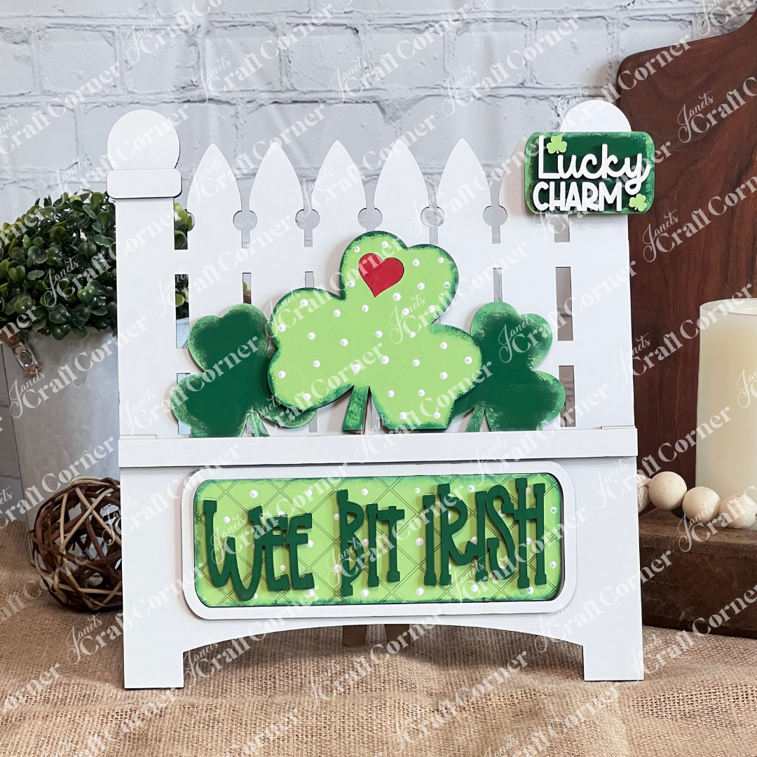 The Wee Bit Irish Interchangeable Insert by Janet's Craft Corner is a decorative wooden piece showing Irish pride with a white picket fence and green shamrock adorned with a heart, featuring "Lucky Charm" and "Wee Bit Irish" signs in green and white, set among candles and a plant.