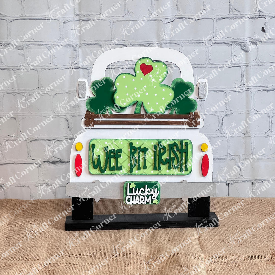 The Wee Bit Irish Interchangeable Insert by Janet's Craft Corner is a decorative white truck cutout featuring green shamrocks and "Wee Bit Irish" text. It sports a "Lucky Charm" license plate and rests on burlap with a brick wall backdrop, perfect for home decor.