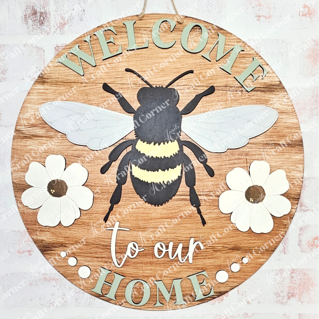 Janet's Craft Corner presents the Welcome Home Bees & Blooms Door Hanger, featuring a bee illustration and two white flowers on a light wood background. The phrase "Welcome to our Home" encircles the bee, perfect for DIY projects and enhancing your entryway decor.