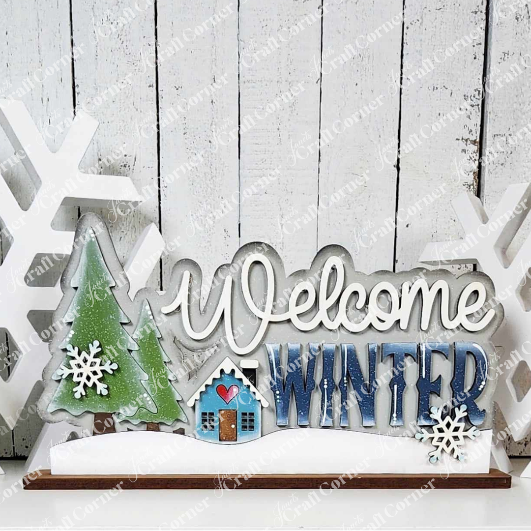 Introducing the "Welcome Winter Word Art" by Janet's Craft Corner — a charming DIY craft kit that enhances your home décor. This hand-painted wooden winter sign features the phrase "Welcome Winter," accompanied by two green trees, a blue house with a red heart, and snowflake decorations, all set against a rustic white wooden wall. Perfect for adding a touch of seasonal warmth to your space!