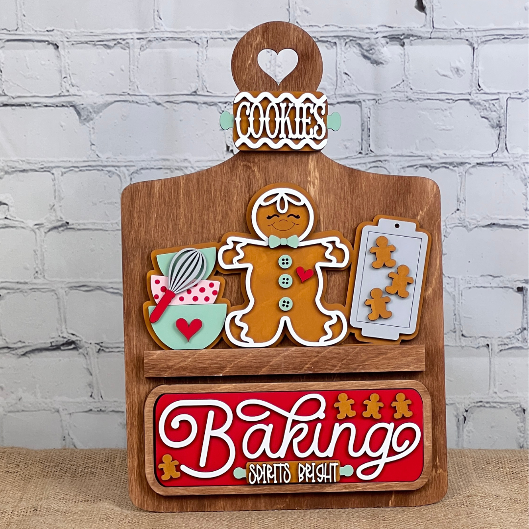 The Breadboard Interchangeable Base with Insert by Janet's Craft Corner features a DIY wooden baking theme. It boasts a gingerbread man, utensils in a bowl, and a cookie tray against a white brick wall. A red "Baking Spirits Bright" sign and heart cutout labeled "Cookies" enrich your home decor.