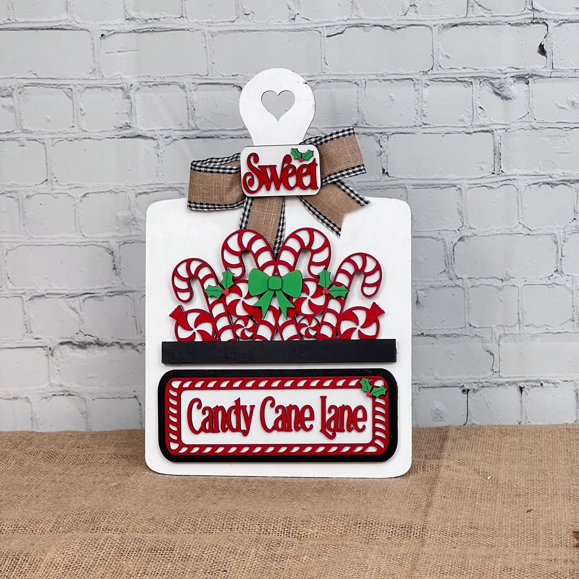 Janet's Craft Corner's Breadboard Interchangeable Base with Insert features a DIY board with "Sweet" and "Candy Cane Lane," highlighting red and white candy canes, a green bow, and heart cutout. Perfectly set on a burlap surface against a white brick wall for unique home decor inspiration.