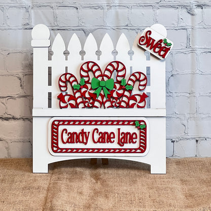 Janet's Craft Corner offers a "Garden Fence with Insert" DIY decor kit. This set includes a festive white picket fence decoration featuring interchangeable inserts like red and white candy canes and green holly, labeled "Candy Cane Lane" in red letters with a small "Sweet" sign on top, against a charming textured wall background.