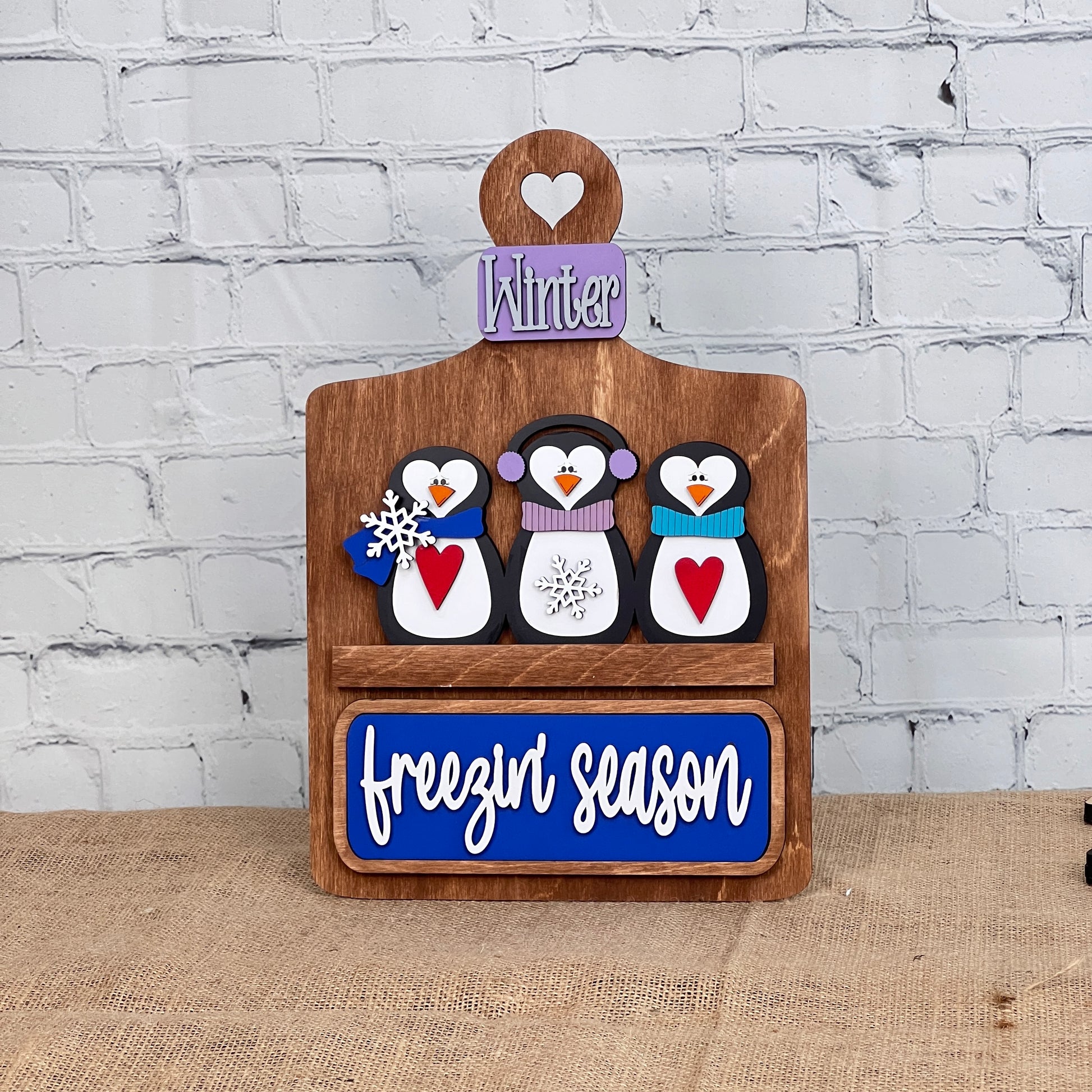 A charming DIY home decor project from Janet's Craft Corner, this wooden breadboard base insert features three penguins with earmuffs and scarves, each holding a red heart. The middle penguin has a snowflake, topped by a "Winter" sign and "freezin' season" on a blue plaque, set against a white brick wall.