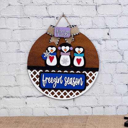 Janet's Craft Corner offers a DIY Kit with an interchangeable door hanger base and insert. It features a wooden round sign with penguins in hats and scarves, a "Winter" banner, and "freezin' season" text on blue. A white lattice pattern completes this charming home décor piece.
.