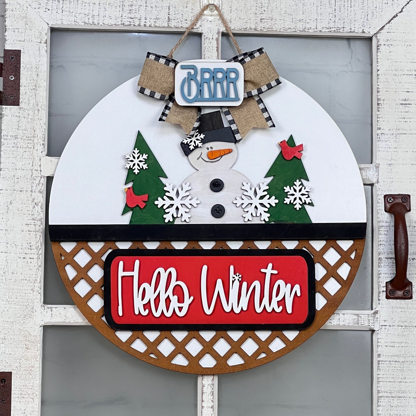 Janet's Craft Corner offers a DIY craft kit featuring a round winter wreath door hanger. This interchangeable home décor showcases a snowman, trees, snowflakes on white, topped with "BRRR" and red "Hello Winter" signs. It includes a black-and-white ribbon for the perfect finish.