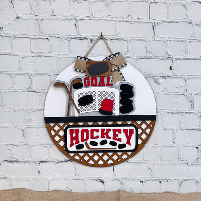 Janet's Craft Corner presents a DIY kit featuring a rustic round wall decor with "GOAL" and "HOCKEY" text, complete with a burlap bow for easy assembly. The interchangeable design includes a hockey net, sticks, puck, and skates, perfect for stylish home décor on a white brick wall.