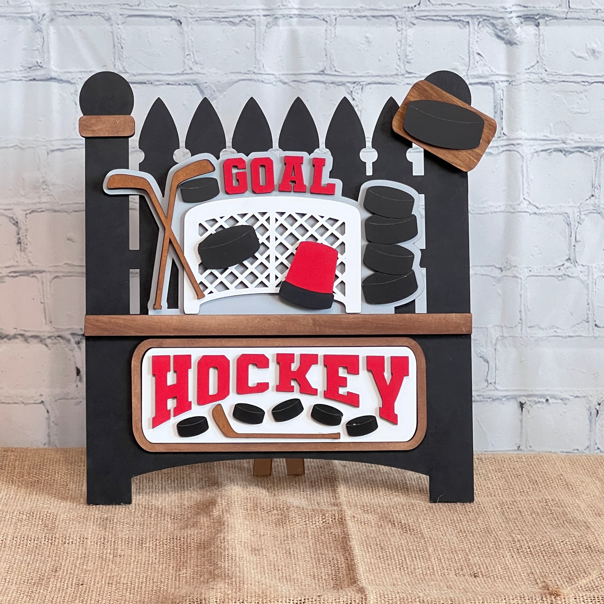 The Garden Fence with Insert from Janet's Craft Corner is a DIY interchangeable decor kit featuring a hockey-themed insert with bold red "Goal" and "Hockey," hockey sticks, pucks, and net on brick wall background. Ideal for craft enthusiasts, it rests on a rustic burlap surface.