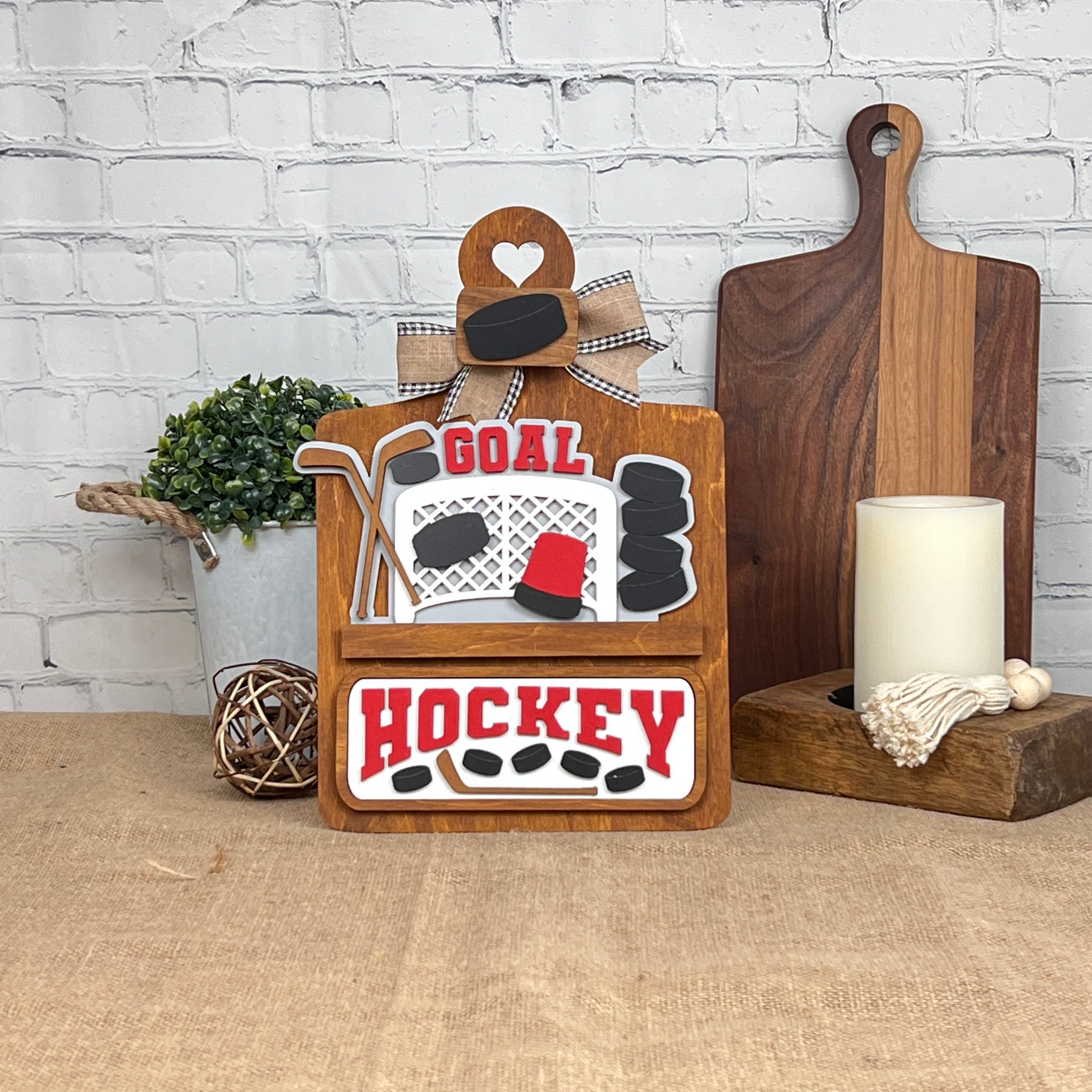 Janet's Craft Corner offers a Breadboard Interchangeable Base with Insert, a DIY kit featuring "GOAL" and "HOCKEY" on a decorative plaque with hockey motifs. It's perfect home decor, complete with a bow, and complements any space with accompanying items like a potted plant or candle.