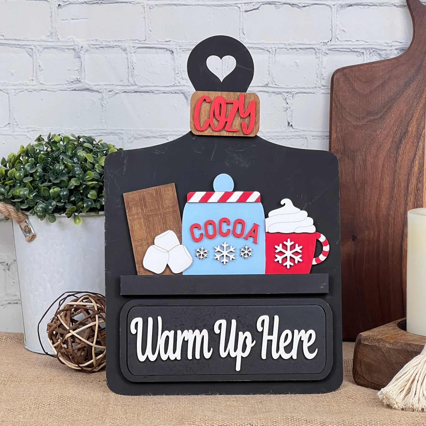 Janet's Craft Corner presents a DIY home decor kit featuring a "Warm Up Here" sign with cocoa illustrations. The included insert has decorative elements like whipped cream, marshmallows, a red "Cozy" label, and background details resembling a plant and candle on wooden boards.