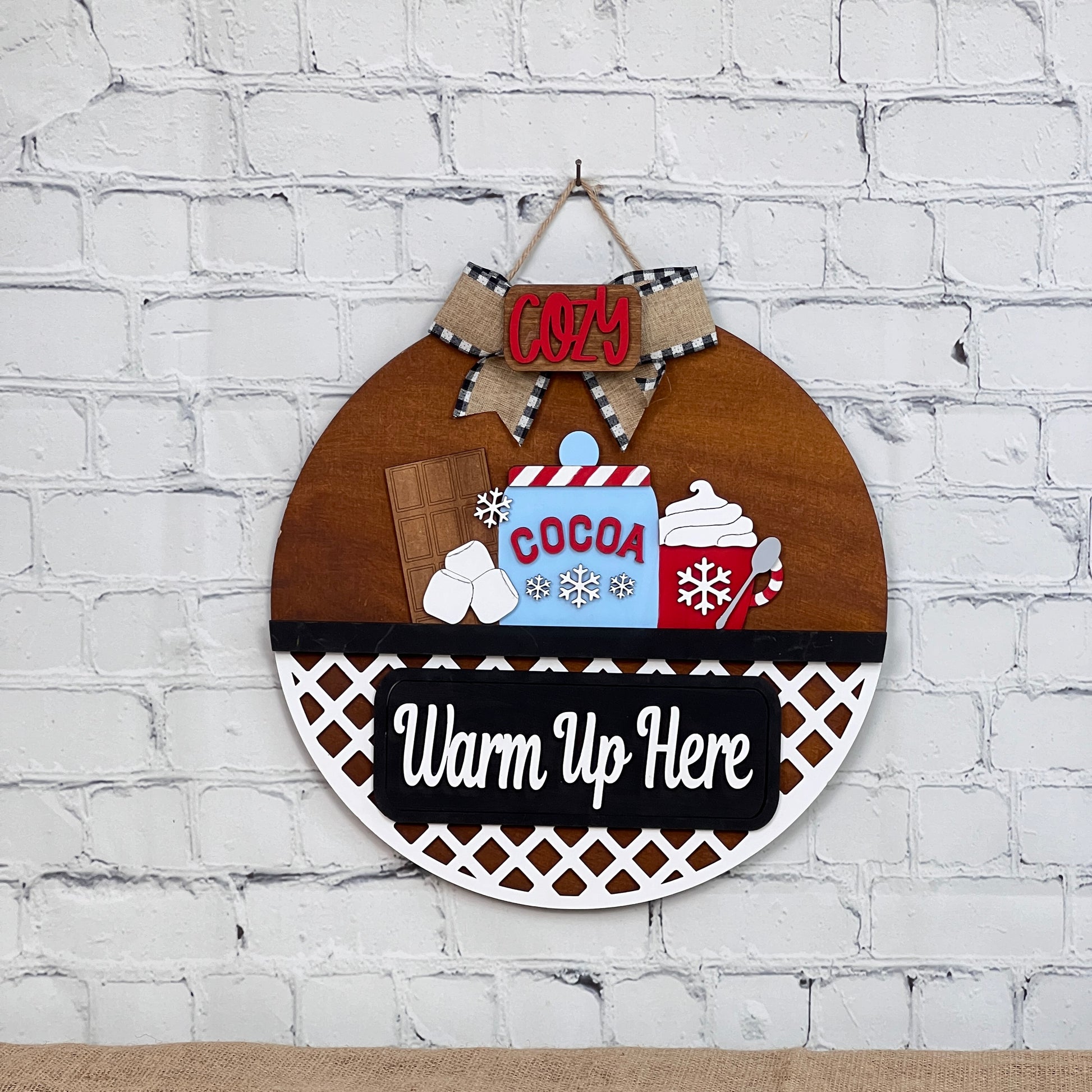 A Janet's Craft Corner DIY kit showcases a round wooden sign against a white brick wall, featuring a cocoa mug with candy canes, marshmallows, whipped cream, and the words "Cozy" and "Warm Up Here," enhanced by an elegant lattice design.