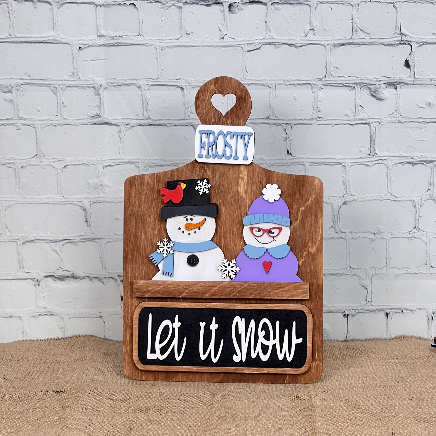 The Breadboard Interchangeable Base with Insert from Janet's Craft Corner is a charming DIY home decor piece featuring a wooden sign with heart cutout, "Frosty," two snowmen (one in a black hat, the other in earmuffs and glasses), and "Let it Snow" in vintage-style script.