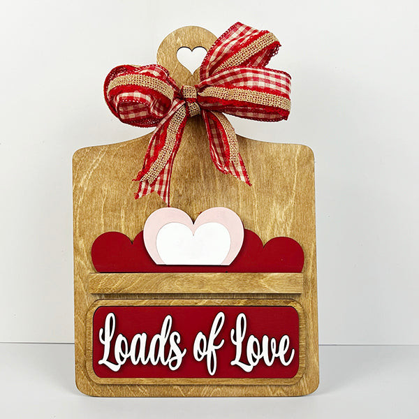 The Breadboard Interchangeable Base with Insert by Janet's Craft Corner features a wooden board with heart cutouts and a red-and-white plaid ribbon. "Loads of Love" is written above two hearts, perfect for creative DIY home decor and ideal for interchangeable accessories.