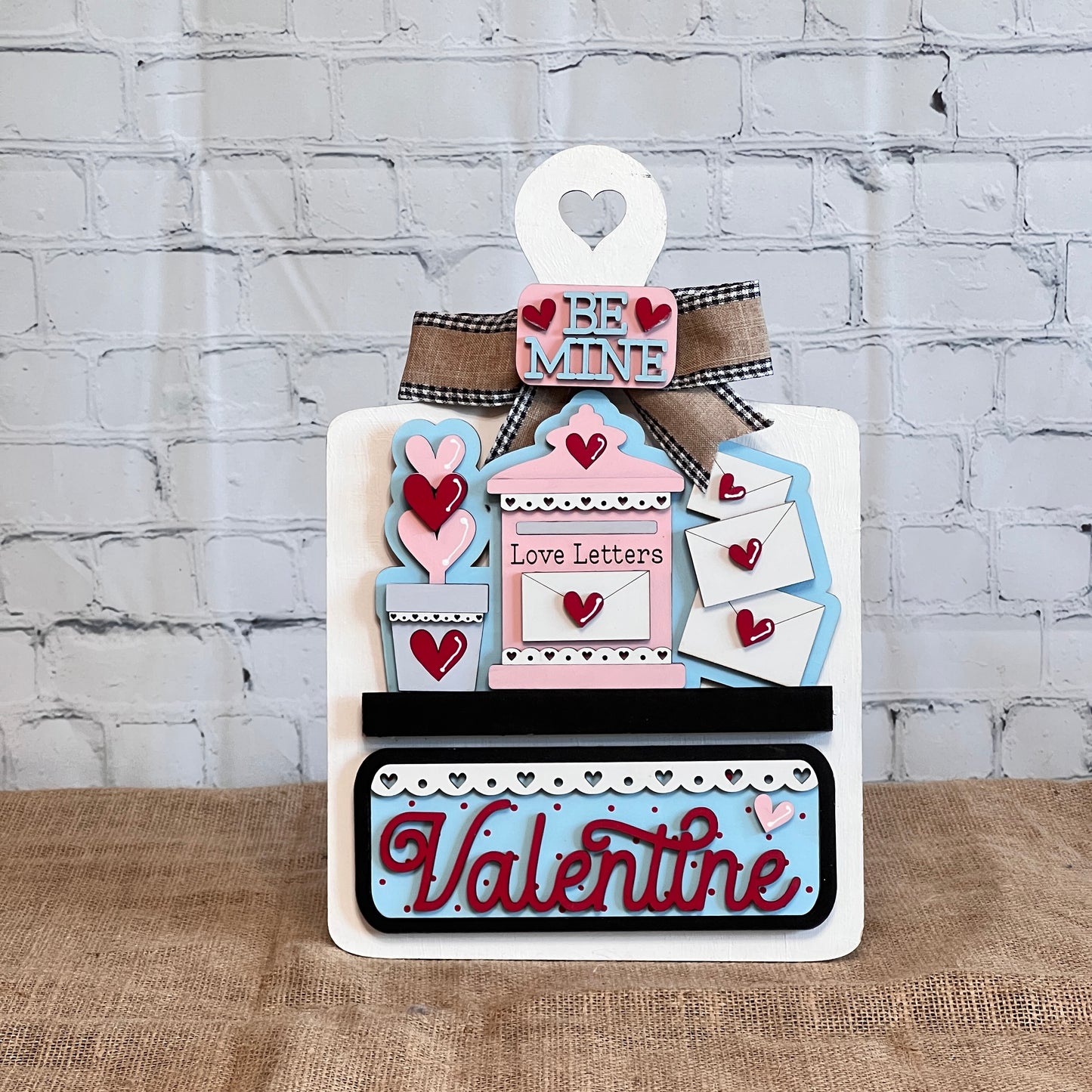 Janet's Craft Corner offers a DIY Valentine's Day decor kit with a breadboard base and insert. It features phrases like "Be Mine" and "Love Letters," adorned with hearts. With pink, blue, and red accents, it enhances home decor beautifully against a brick wall background.