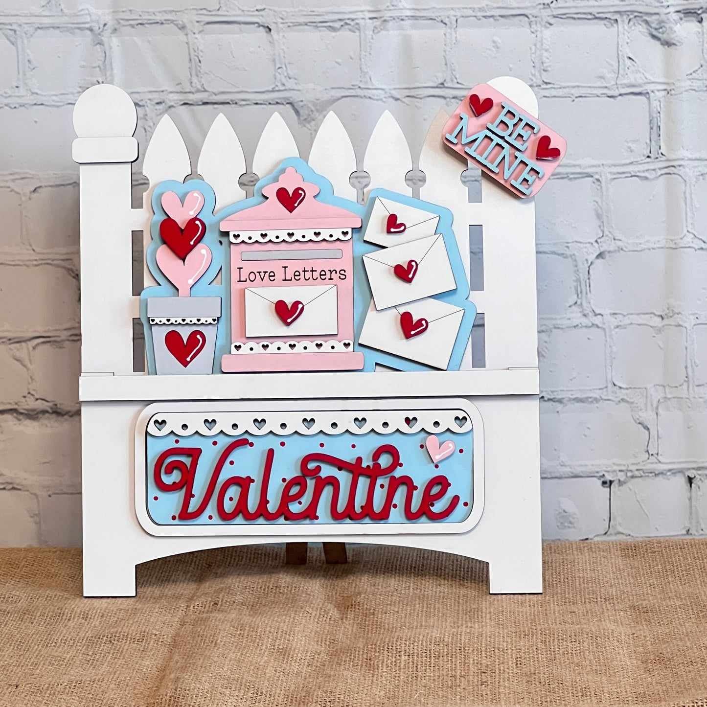 Janet's Craft Corner offers a Garden Fence with Insert DIY kit, featuring a white picket fence with interchangeable inserts. It includes a "Love Letters" mailbox, heart envelopes, and a "Be Mine" sign. The word "Valentine" is in red on a blue background below.