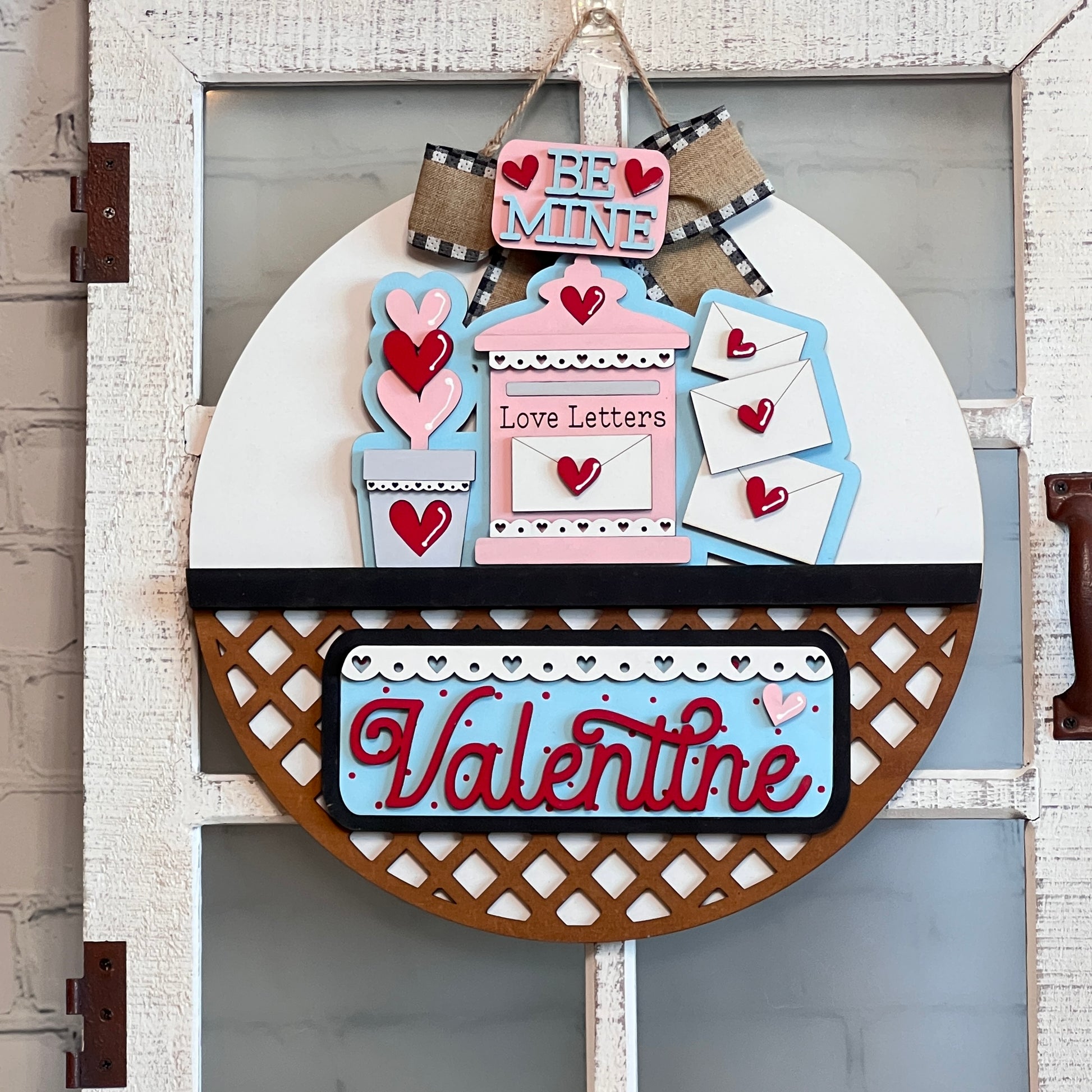 Janet's Craft Corner offers a DIY interchangeable home decor kit featuring a round door hanger with cursive phrases "Be Mine," "Love Letters," and "Valentine" on a rustic white background. It includes pink and blue envelopes, heart accents, bows, lattice design—ideal for decorative flair.