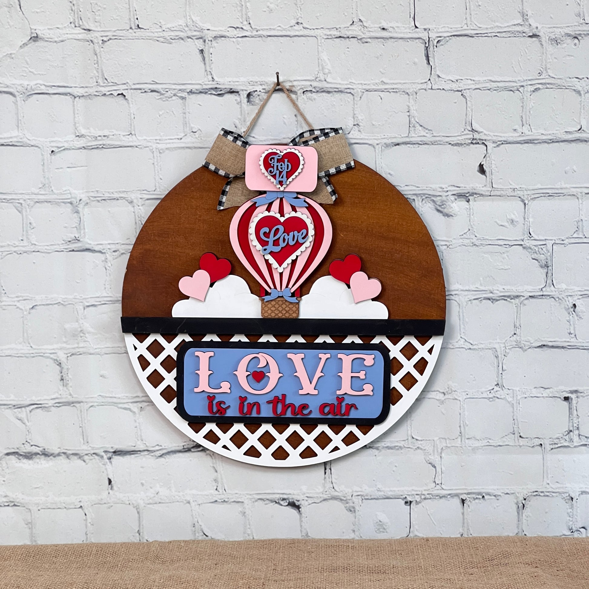 Janet's Craft Corner offers a DIY set featuring a round door hanger with a hot air balloon and "Love is in the air" design. It hangs on a white brick wall with a bow, adding charm to your home year-round.